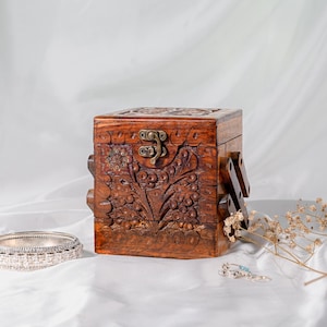 Könnte beinhalten: Eine quadratische Holzschmuckschatulle mit aufwendigen Blumenschnitzereien und einem Messingverschluss. Die Schatulle hat eine warme braune Farbe und einen Klappdeckel. Ein silbernes Armband und kleine Ohrringe liegen neben der Schatulle.