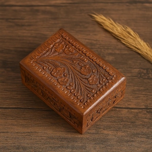 May include: A rectangular wooden box with intricate floral carvings on the lid and sides. The box is a warm brown color and appears to be made of a solid wood. The carvings feature detailed flowers and leaves, creating a decorative and textured surface.