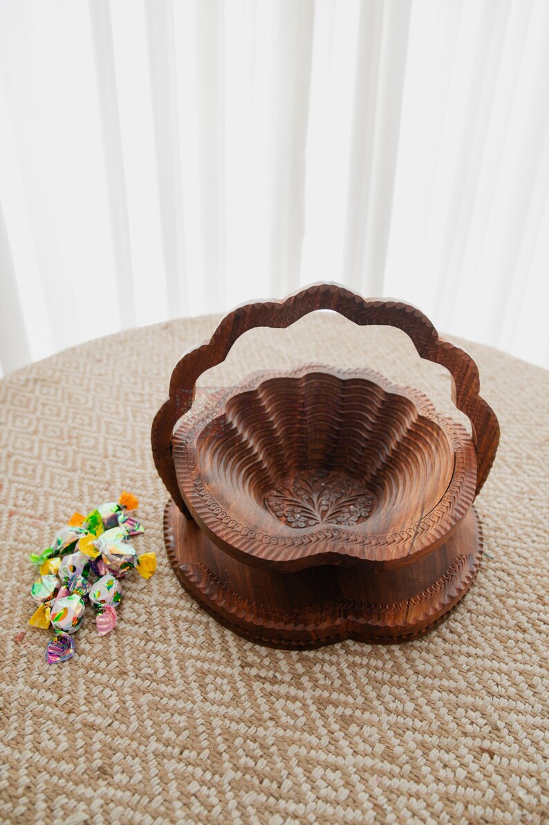 May include: A carved wooden fruit basket with a handle. The basket is made of dark brown wood and has a scalloped edge. The interior is carved with a floral design. A small pile of wrapped candies sits to the left of the basket.