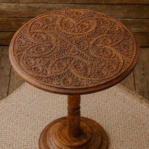 Puede incluir: Una mesa auxiliar redonda de madera con intrincadas tallas florales en la superficie. La mesa tiene una base de pedestal y está teñida de un color marrón cálido. La mesa tiene aproximadamente 61 cm de diámetro.