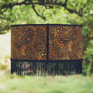 Può includere: Paralume cilindrico con motivo leopardato nei toni del marrone e del nero. Il paralume è rifinito con tessuto nero e una frangia di nappe nere sul fondo. Il paralume è appeso a un filo sottile.