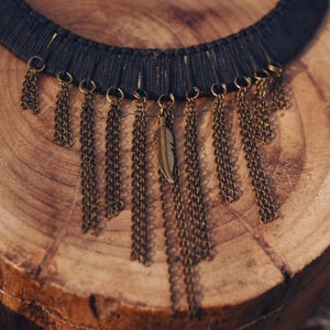 May include: A dark gray choker necklace with dangling bronze-colored chains and a single feather pendant. The necklace is displayed on a textured, light brown wooden surface, showcasing a bohemian style.