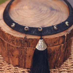 May include: A black choker necklace with a decorative tassel. The necklace features a woven band adorned with small, round, gold-toned embellishments. The tassel is long and made of black threads, with a gold-toned, patterned metal cap.