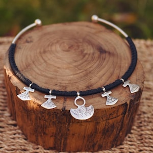 May include: A black choker necklace with silver-toned pendants. The pendants include axe and crescent moon shapes. The necklace is displayed on a wooden surface, with a natural background.