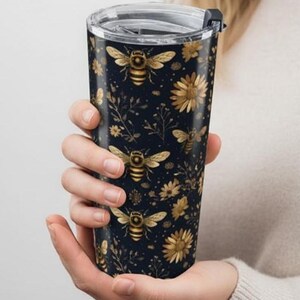 Puede incluir: Un vaso de viaje negro con un patrón floral y de abejas doradas. El vaso tiene una tapa transparente. El diseño presenta abejas, flores y follaje dorados sobre un fondo oscuro.