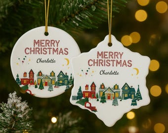 Peut inclure: Deux décorations de Noël blanches avec une ficelle dorée. L'une est ronde, l'autre en forme de flocon de neige. Les deux portent "JOYEUX NOËL" en rouge, le nom "Charlotte" et une scène de village.