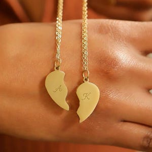 Puede incluir: Dos collares dorados con colgantes en forma de corazón. Uno tiene la letra "A", el otro "K". Los colgantes están divididos, sugiriendo amistad o relación.