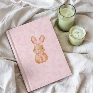 Puede incluir: Un diario rosa con un estampado floral y una ilustración de acuarela de un conejito con un lazo. Una vela verde en un tarro de cristal y una pequeña taza de matcha están junto al diario.