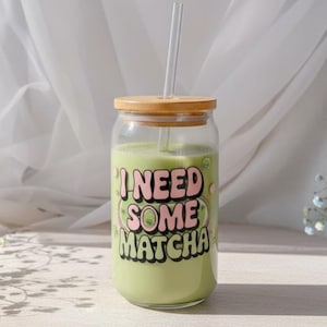 Puede incluir: Un vaso de vidrio transparente lleno de líquido verde, una pajita y una tapa de madera. El vaso tiene el texto "I NEED SOME MATCHA" en rosa y verde. El vaso está sobre una superficie clara.