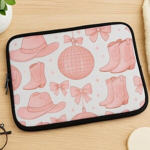 May include: A laptop sleeve with a repeating pattern of pink cowboy hats, boots, bows, and disco balls on a white background. The sleeve has a black zipper and trim.