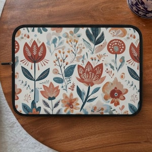 Puede incluir: Una funda rectangular para portátil con un estampado floral. El diseño presenta flores estilizadas en tonos óxido, melocotón y azul sobre un fondo crema. La funda tiene una cremallera y ribete negros, y está sobre una superficie de madera.