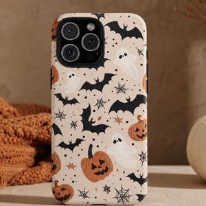 Puede incluir: Una funda para teléfono con fondo beige claro que presenta un patrón repetitivo de murciélagos negros, fantasmas blancos, calabazas naranjas y telarañas negras. La funda tiene un recorte rectangular para un sistema de cámara de tres lentes.