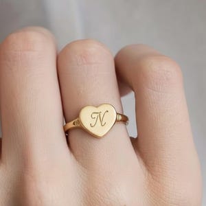Puede incluir: Anillo dorado con una cara en forma de corazón. El anillo presenta la letra "N" grabada en el corazón. La banda tiene un diseño sencillo. El anillo se muestra en un dedo.