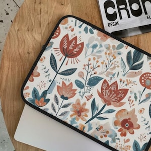 Può includere: Custodia per laptop con motivo floreale nei toni dell'arancione, verde acqua e bianco. Il design presenta fiori e foglie stilizzate su sfondo crema, con bordo nero. La custodia è su un tavolo di legno.