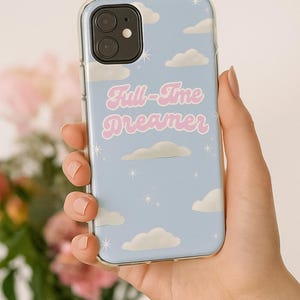 Puede incluir: Funda de teléfono azul claro con un diseño de nubes y estrellas. La funda presenta el texto "Full-Time Dreamer" en rosa. La funda está sujeta en una mano.