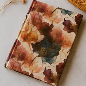 Puede incluir: Un diario cerrado con un diseño floral. La portada presenta un patrón de flores estilizadas en tonos burdeos, melocotón y verde azulado sobre un fondo crema. El diario es rectangular y parece estar hecho de un material liso.