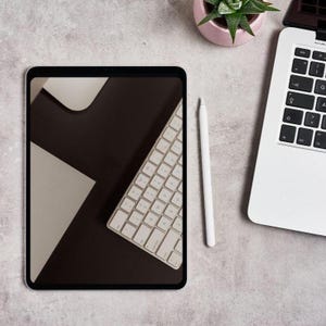 May include: A digital tablet with a keyboard displayed on the screen, a white stylus, and a laptop are arranged on a light gray surface. A small pink pot with a green plant is also present, creating a modern workspace aesthetic.