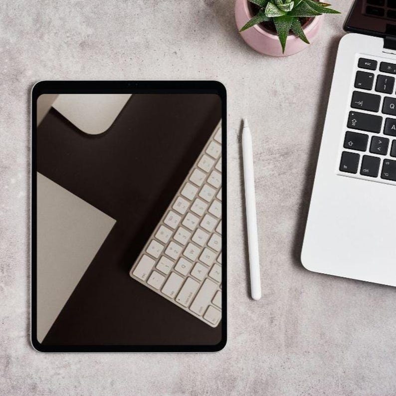 May include: A digital tablet with a keyboard displayed on the screen, a white stylus, and a laptop are arranged on a light gray surface. A small pink pot with a green plant is also present, creating a modern workspace aesthetic.