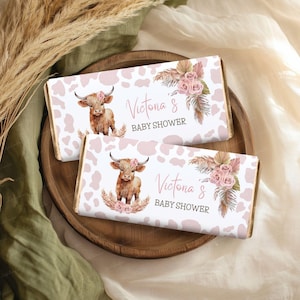 Può includere: Due incarti rettangolari per barrette di cioccolato con uno sfondo a macchie di mucca rosa chiaro. Ogni incarto presenta un'illustrazione di una mucca highland marrone, rose rosa, foglie di palma essiccate e il testo Victoria's BABY SHOWER.