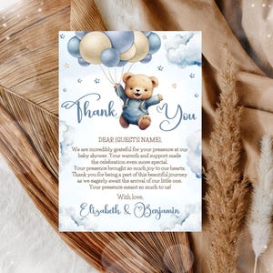 Op de afbeelding: Een bedankkaart voor een babyshower met een aquarel illustratie. Een teddybeer in een blauwe outfit zweeft met ballonnen. De kaart heeft de woorden "Thank You" en een boodschap van dankbaarheid, met de namen Elizabeth & Benjamin.