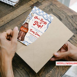 Puede incluir: Una invitaci&oacute;n para baby shower con una ilustraci&oacute;n de botella de cola de estilo vintage. La invitaci&oacute;n dice "She's Ready to Pop!" en escritura roja, con detalles del evento para Emily Johns. La invitaci&oacute;n est&aacute; en un sobre beige.