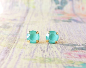 May include: A pair of small, round, turquoise blue earrings with gold settings. The earrings are on a light wood background with a floral pattern.