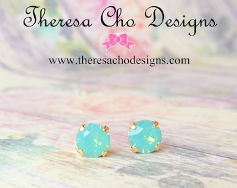 May include: A pair of small, round, light blue earrings with gold settings. The earrings have a sparkly, iridescent finish.