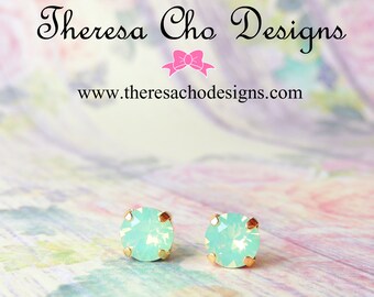 May include: A pair of gold stud earrings with light green opal stones. The earrings are on a light pink and white background.