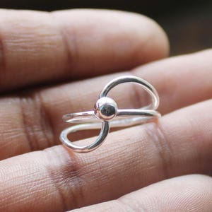 May include: A silver ring with a unique design. The ring features a small sphere at the center, with curved bands extending around it. The ring is made of a shiny, reflective metal, and is shown against a blurred background.
