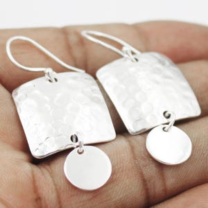 以下が含まれることがあります： 銀色のイヤリングのペア。各イヤリングは、小さな丸いチャームが下にぶら下がっている、ハンマーで叩かれた正方形のトップが特徴です。イヤリングはシンプルなフックデザインです。金属は明るく反射する仕上がりです。