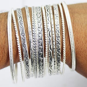 May include: A collection of silver bangles with various decorative patterns. The bangles include floral, leaf, and rope designs. The bangles are displayed on a person's arm, showcasing the different textures and styles of the jewelry.