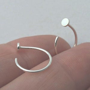 Op de afbeelding: Twee zilveren neusringen, elk bestaande uit een dunne, gebogen metalen draad die aan één uiteinde eindigt in een kleine, platte, ronde schijf. De metalen sieraden hebben een gepolijst, glad en reflecterend oppervlak met een eenvoudig ontwerp.