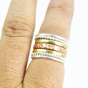 Puede incluir: Un anillo multibanda con bandas plateadas, doradas y cobrizas. El anillo presenta una banda plateada trenzada en la parte superior e inferior, con bandas doradas y cobrizas en el medio. La banda de cobre tiene un patrón texturizado.