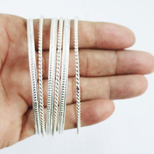 Puede incluir: Un conjunto de pulseras de plata con un diseño de cuerda trenzada. Las pulseras delgadas tienen una superficie brillante y reflectante. Las joyas se exhiben sobre un fondo neutro.