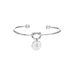 Puede incluir: Una pulsera de brazalete plateada con un diseño de nudo en el centro y un pequeño dije redondo. El dije tiene grabada la letra "S". La pulsera tiene un diseño simple y elegante.