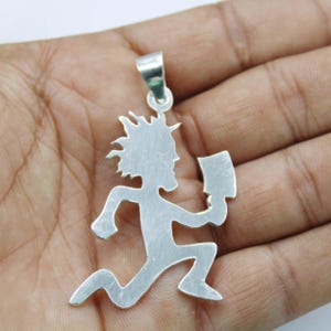 May include: A silver-coloured metal pendant shaped like a running figure with spiky hair, holding a cleaver in one hand. The pendant features a smooth, flat surface and includes a small metal loop at the top for attachment to a chain or cord.
