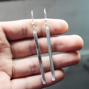 以下が含まれることがあります： シルバーのバーピアス。各ピアスは、小さなフックから吊り下げられた細長い長方形のバーが特徴です。ピアスは光沢のある反射性の金属で作られています。