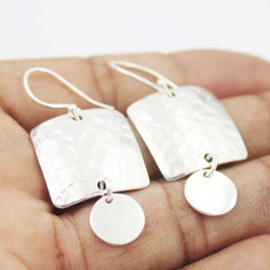 Peut inclure: Une paire de boucles d'oreilles pendantes argentées avec un pendentif carré à la surface martelée et texturée. Chaque pendentif carré est fixé à un petit disque circulaire argenté lisse qui pend au bord inférieur du carré.