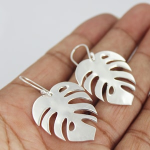 May include: A pair of silver Monstera leaf earrings. The earrings are shaped like the iconic split-leaf philodendron plant, with cut-out sections. The earrings are shown against a neutral background.