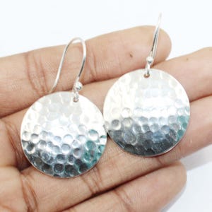 Può includere: Un paio di orecchini a disco martellati in argento. Gli orecchini hanno una superficie strutturata con un design martellato. Sono rotondi e pendono da un semplice gancio. Lo sfondo è chiaro.