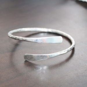 Op de afbeelding: Een zilverkleurige open armband met een getextureerde, gehamerde afwerking langs de band en gladde, platte, afgeronde uiteinden. Het metaal heeft een reflecterend oppervlak met zichtbare inkepingen door het hamerproces in het ontwerp.
