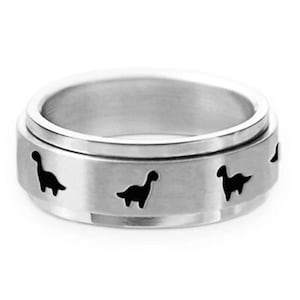 Op de afbeelding: Een zilverkleurige spinner ring met een geborstelde metalen afwerking. De ring heeft uitgesneden silhouetten van dinosaurussen rond de band. De ring is ontworpen om te draaien, wat een tactiele ervaring biedt.