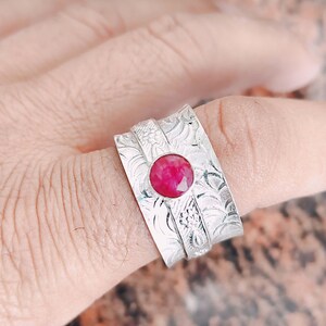 May include: A silver ring with a central, round, ruby-red gemstone. The ring has an engraved floral pattern and a wide band. The gemstone is the focal point, set in a raised bezel. The ring is on a finger.