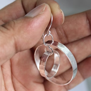 Puede incluir: Pendientes colgantes plateados con un diseño único e entrelazado. Los pendientes presentan dos aros conectados, uno con una textura martillada y el otro con un acabado liso y pulido. Los pendientes están suspendidos de un gancho simple.