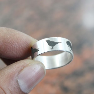 May include: A silver ring with a repeating pattern of black bird silhouettes. The ring is held between fingers, showcasing the polished metal and the detailed bird designs. The ring is a piece of jewelry.
