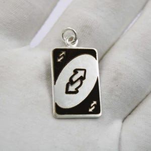 Puede incluir: Un colgante plateado con forma de carta de reversa del popular juego de cartas. El colgante tiene un borde negro y presenta un óvalo blanco con dos flechas curvas. El colgante es rectangular con esquinas redondeadas.