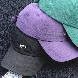May include: Three baseball caps in green, purple, and black are stacked. The black cap has the word "Nice" embroidered on the front. The caps have a curved peak and a fabric strap at the back. The caps are made of a lightweight material.