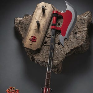 May include: A red and silver battle axe with a wooden handle, displayed on a textured wooden base. A wooden plaque with three pegs and a red emblem is mounted to the left. The axe blade is silver with a red accent.
