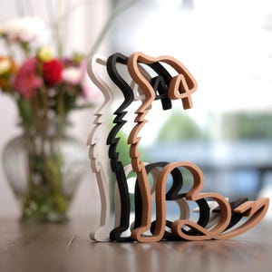K&ouml;nnte beinhalten: Ein mehrschichtiges Holzskulptur eines Hundes in Wei&szlig;, Schwarz und Braun. Der Hund sitzt, wobei jede Schicht leicht versetzt ist, was einen 3D-Effekt erzeugt. Die Skulptur steht auf einer Holzoberfl&auml;che. Die Skulptur ist etwa 20 cm hoch.