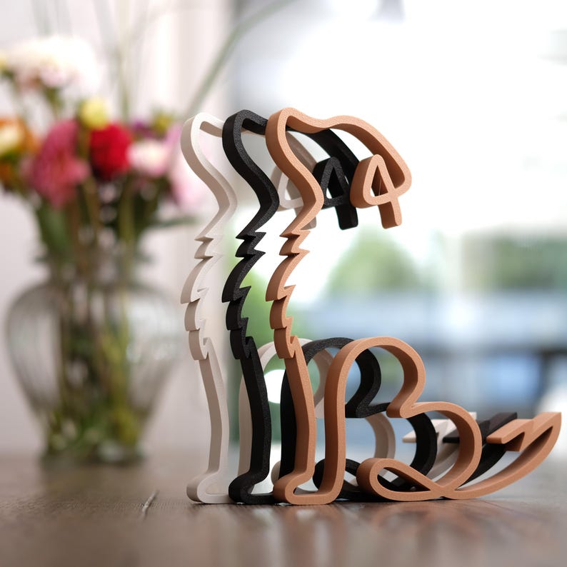 K&ouml;nnte beinhalten: Ein mehrschichtiges Holzskulptur eines Hundes in Wei&szlig;, Schwarz und Braun. Der Hund sitzt, wobei jede Schicht leicht versetzt ist, was einen 3D-Effekt erzeugt. Die Skulptur steht auf einer Holzoberfl&auml;che. Die Skulptur ist etwa 20 cm hoch.